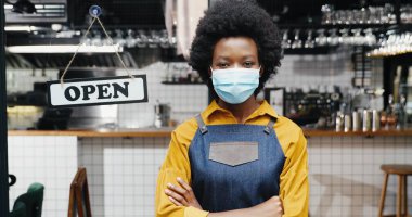 Önlüklü ve tıbbi hizmetli güzel Afro-Amerikan bayan garsonun portresi. Kafe girişinde masası açık bir şekilde kameraya bakıyor. Güzel barikat kapısı açık duruyor. Tekrar aç.