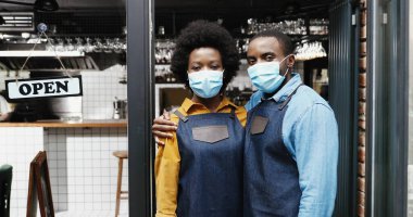 Afro-Amerikalı önlüklü ve tıbbi maskeli birkaç neşeli barmenin portresi kameraya mutlu bir şekilde gülümsüyor ve kafe girişindeki açık masada sarılıyorlar. Bar kapısındaki mutlu garson ve garson. Yeniden Aç.