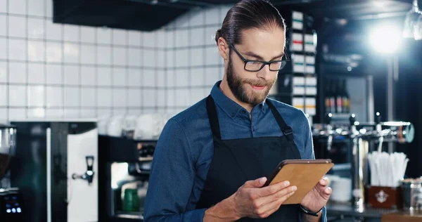 ガラスやエプロンで笑顔とタブレットデバイスを使用してバーでチャットするときにハンサムな若い白人男性ウエイター 男バーテンダータップ スクロールし キッチンでガジェットのコンピュータ上のテキストメッセージ — ストック写真