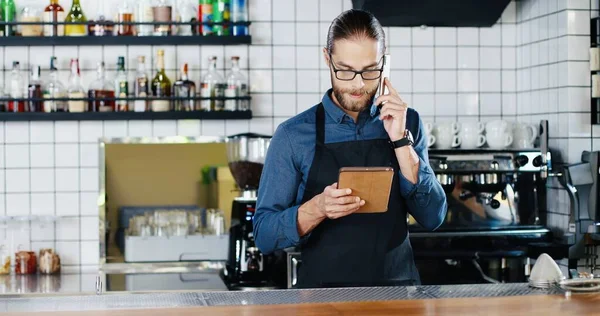 Handsome young male Caucasian bartender talking on mobile phone and holding tablet device at counter in bar. man barrista in glasses and aprone speaking on cellphone and tapping on gadget.