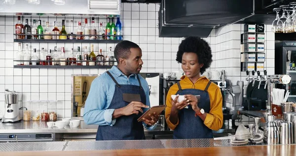 Афроамериканец красивый молодой бармен в фартуке держит планшетное устройство и барристу, пишущую список заказов или делающую покупки на кухне бара. Официант и официантка составляющие меню для кафе — стоковое фото