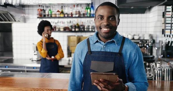 Kafede tablet kullanan ve kameraya gülümseyen Afrikalı Amerikalı erkek barmenlerin portresi. Yakışıklı garson. Cihazla internete bağlanıp kayıyor. Garson arka planda cep telefonuyla konuşuyor.. — Stok fotoğraf