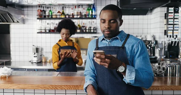 Manliga afroamerikanska barmen använder smartphone i café och SMS. Snygg man servitör knacka och bläddra online på telefon. Servitris med surfplatta på bakgrunden. Kvinnliga hållande prylar. — Stockfoto