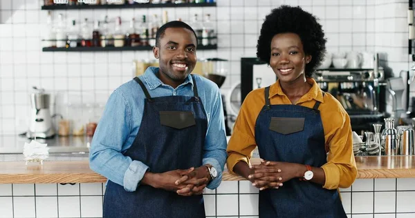 Portrait of African American cheerful couple of bartenders in aprons looking at each other and smiling to camera. Happy waiter and waitress posing in cafe. Joyful man and woman, barristas at work.