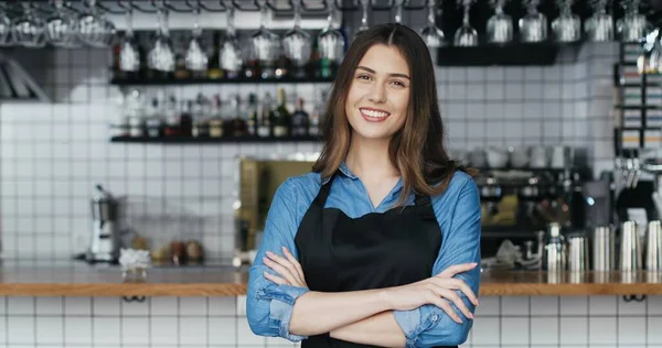 Porträtt av vit ung glad vacker kvinna barrista i förkläde står vid disken i baren, ler mot kameran och korsar händer. Servitris poserar i café med dryckesutrustning på bakgrunden. — Stockfoto