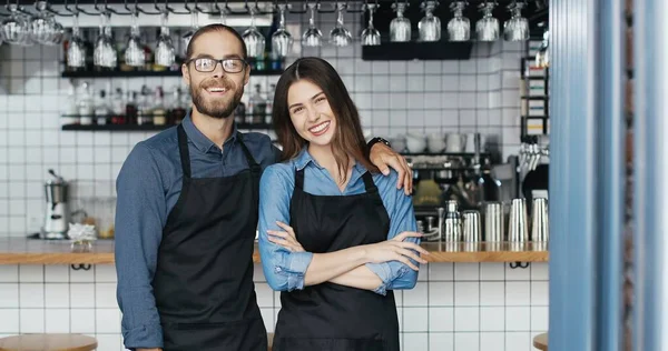 Porträtt av vita glada par bartenders i förkläden kramar varandra och ler glatt mot kameran. Glad servitör och servitris i kramar på caféet. En glad man som omfamnar kvinnan. Barriärer. — Stockfoto