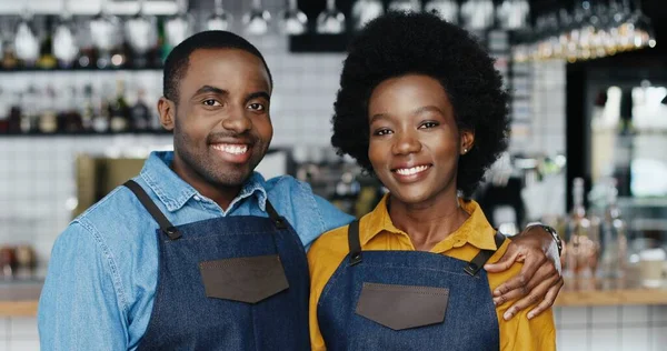 Porträtt av afroamerikanska glada par bartenders i förkläden kramar varandra och ler glatt mot kameran. Glad servitör och servitris i kramar på caféet. En glad man som omfamnar kvinnan. Barriärer. — Stockfoto