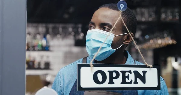 Cameriere afroamericano in maschera medica fasciatoio sulla porta - chiuso-aperto al bar inmmorning. Bar di apertura maschile in anticipo e sorridente. Allegro venditore girando bordo e apre negozio. Misure pandemiche — Foto Stock