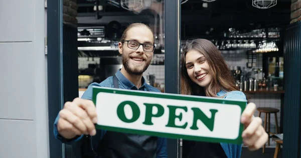 Porträtt av vita glada par bartenders i förkläden ler gladeligen mot kameran och håller bordet öppet vid caféentrén. Glad servitör och servitris vid bardörren med öppet bord. Glädjande man kvinna. — Stockfoto