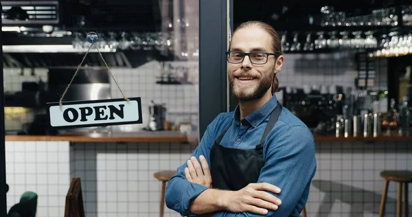 Kafe girişindeki masası açık kameraya gülümseyen önlüklü ve gözlüklü yakışıklı, genç, mutlu adam garson portresi. Neşeli, gülümseyen, barın kapısında duran, tahtası açık erkek bariyer. — Stok fotoğraf