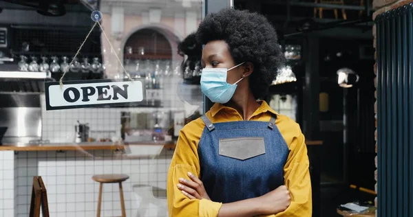 Retrato de bela jovem garçonete afro-americana em avental e médico olhando para câmera com mesa aberta na entrada do café ao ar livre. Bastante barrista em pé na porta do bar com placa aberta Reabrir — Fotografia de Stock