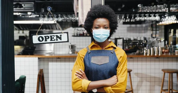 Retrato de bela jovem garçonete afro-americana em avental e médico olhando para câmera com mesa aberta na entrada do café ao ar livre. Bastante barrista em pé na porta do bar com placa aberta Reabrir — Fotografia de Stock