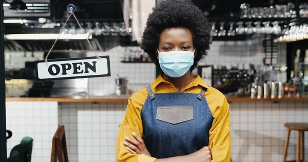 Retrato de la hermosa joven afroamericana camarera en delantal y médico mirando a la cámara con mesa abierta en la entrada de la cafetería al aire libre. Bastante abogado de pie en la puerta del bar con tablero abierto reabrir —  Fotos de Stock