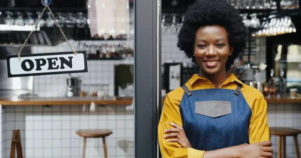 Porträtt av vacker afroamerikansk ung lycklig kvinna servitris i förkläde ler mot kameran med bord öppet vid café entré utomhus. Ganska glad barrista stående vid bardörren med öppen bräda. — Stockfoto