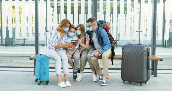 Caucasian happy family in medical masks sitting on bus stop and looking for route on tablet device. Parents with kids and suitcases on wheels waiting for transport and using gadget. Quarantine trip.