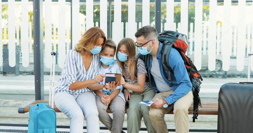 Caucasian happy family in medical masks sitting on bus stop and looking for route on tablet device. Parents with kids and suitcases on wheels waiting for transport and using gadget. Quarantine trip.