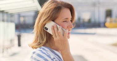 Güneşli bir günde açık havada telefonla konuşan beyaz, güzel, sarışın bir kadın. Otobüs durağındaki cep telefonundan arayan mutlu bir kadın. Bayan cep telefonuyla konuşuyor, nakil için bekliyor..