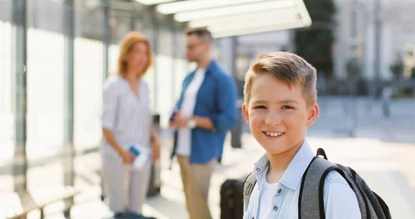 Porträtt av söt vit liten pojke med ryggsäck står utomhus vid busshållplatsen och ler mot kameran. Föräldrar pratar på suddig bakgrund. Familj på resa. Reser. Liten son. — Stockfoto