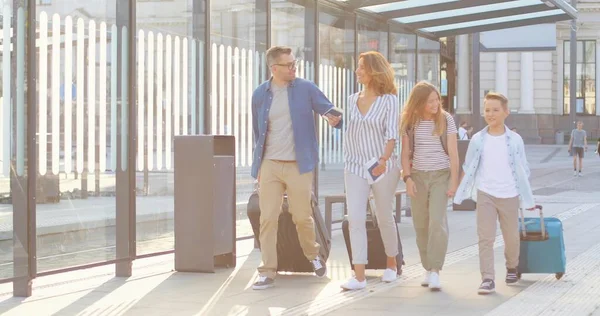 Des parents caucasiens joyeux, deux enfants mignons marchant à l'arrêt de bus, portant des valises sur roues, parlant. Mère père et petite fille et fils à la gare. Bonne famille en vacances en été — Photo