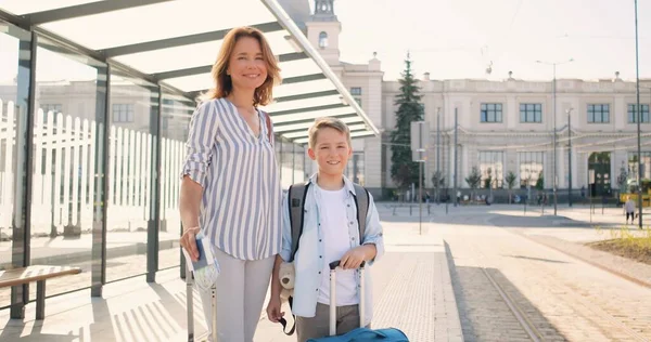 Retrato de joven caucásica hermosa madre y lindo hijo pequeño con maletas en ruedas de pie al aire libre en la parada de autobús y sonriendo a la cámara. Mujer bonita y niño pequeño, turistas en la estación de tren. — Foto de Stock