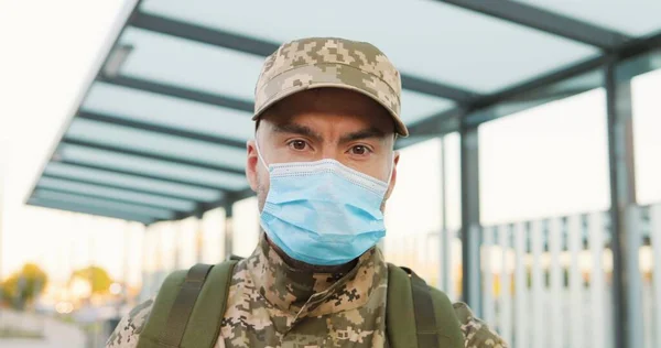 Portrait of serious Caucasian handsome young male soldier in cap and medical mask standing at street. Man militarian looking at camera at train station. Bus stop. Military uniform. Covid-19 pandemic.