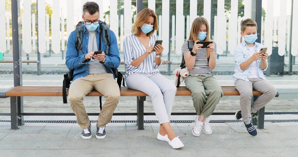 Social distance at bus stop among family members. Caucasian parents with small son and daughter in medical masks sitting on bench, using mobile phones. Tapping scrolling on smarphones. Pandemic trip.