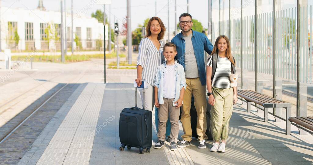 Portrait shot of Caucasian happy family with suitcases standing outdoor at bus sop or train station. Parents with son and daughter travelling in summer. Vacations of children with mother and father.