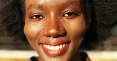 Kameraya bakıp gülümseyen kıvırcık saçlı mutlu Afro-Amerikan kadının genç yüzünü kapatın. Güneş ışığında gülümseyen bir kadın portresi. Açık hava..