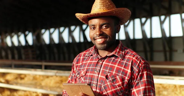 Portret van een Afro-Amerikaanse herder die glimlacht naar de camera in de schuur met vee en tablet-apparaat. Mannelijke boer in schapenstal tikken op gadget. Scrollen op de computer. Surfen online. — Stockfoto