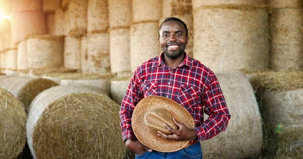 Young handsome African American man farmer standing and smiling in barn with hay barn. Portrait of happy cheerful male shepherd taking off hat and doing greeting gesture in stable. Outdoor. — Stock Photo, Image