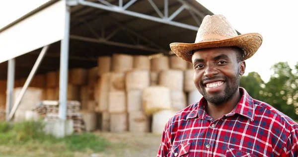 Jeune homme paysan afro-américain beau chapeau debout et souriant avec hangar plein de foin sur fond. Portrait de joyeux berger masculin gai à l'écurie. En plein air. Gros plan. — Photo