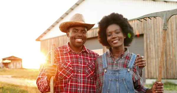 Portrét šťastného veselého afroamerického mladého páru farmářů s vidlemi stojících venku a usmívajících se na kameru ve vesnici. Hezký muž a žena pózují s vidličkami a úsměvy. Farming. — Stock fotografie