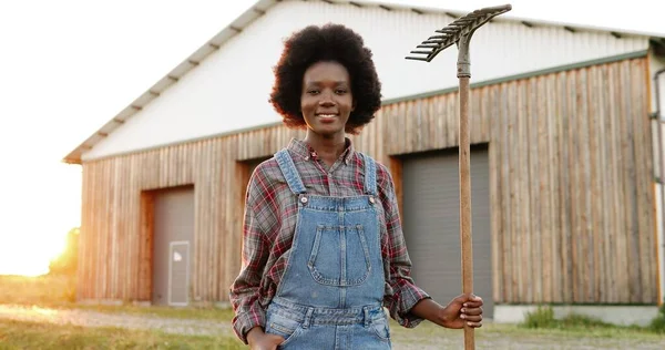 Portrait de belle jeune femme afro-américaine avec fourche agriculteur debout dans le champ à hangar sur fond et souriant à la caméra. Jolie femelle avec sourire et fourchette à la ferme. Concept de campagne. — Photo