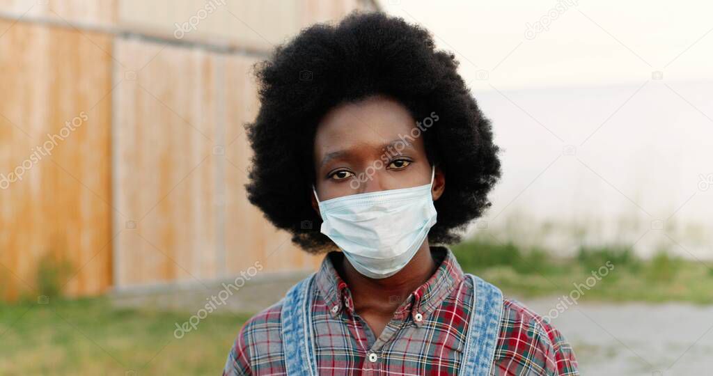 Close up of young African American sad woman with curly hair and in motley shirt outdoors looking at camera. Portrait of female farmer in medical mask standing outside wooden shed. Covid-19 quarantine