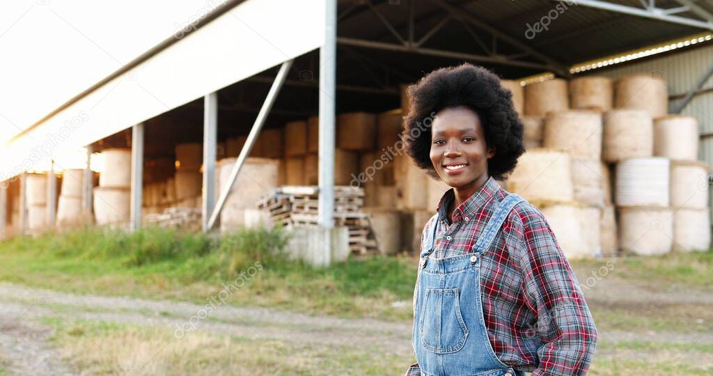 Portrait of beautiful African American young woman farmer standing in field at shed with hay stocks and smiling to camera. Pretty female with smile at farm. Countryside concept.