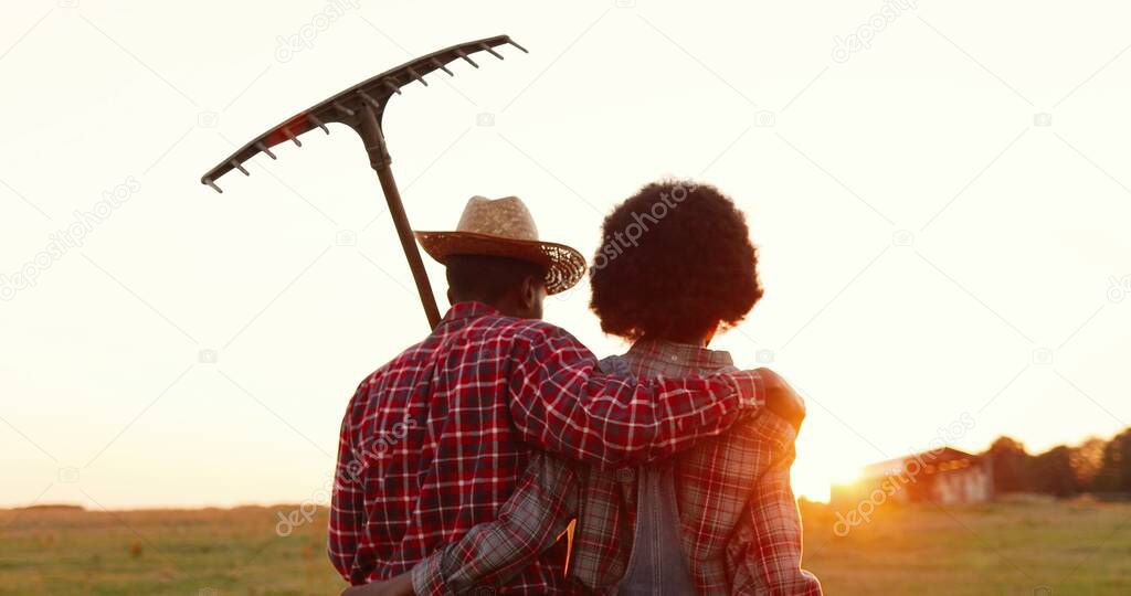 Rear on African American couple of farmers walking in countryside with pitchfork outside and hugging on sunset or sunrise. Back view on man and woman, farm workers strolling to work or coming back.