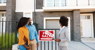 Yeni evimizde kameraya gülümseyen kadın emlakçıyla, maske takmış, evli, Afro-Amerikalı genç bir çift. Ev satıldı. Salgın hastalık konsepti. Konut satın alınıyor.