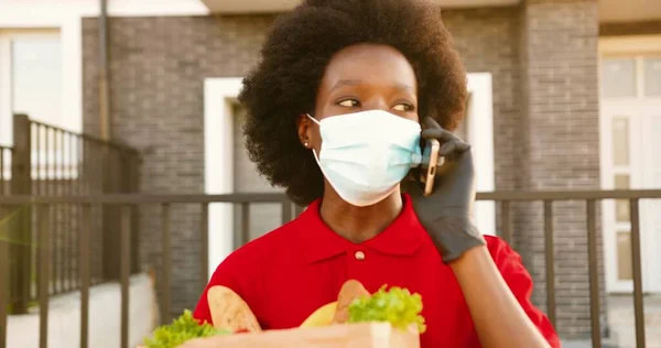 Africano americano jovem mulher de parto em máscara médica e luvas de pé ao ar livre com saco cheio de legumes e falando no celular. Correio feminino a falar no telemóvel. Entregar alimentos. — Fotografia de Stock