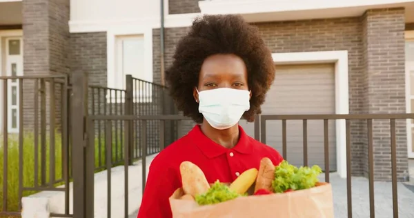 Retrato de una joven mensajera afroamericana en máscara médica y guantes parados en casa en la calle y sosteniendo la bolsa con comida saludable. Trabajadora de reparto con paquete de verduras. Cuarentena. —  Fotos de Stock