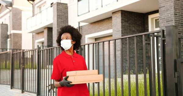 Mensajera joven afroamericana en máscara médica y guantes caminando por la calle y llevando paquetes de cartón. Trabajadora de reparto paseando con cajas de pizza. Concepto de cuarentena. Entrega. —  Fotos de Stock