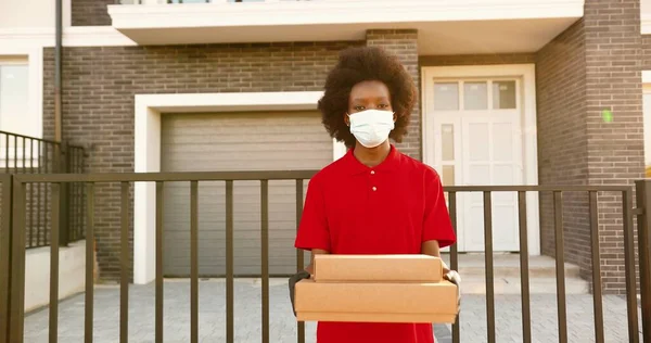 Retrato de la joven mensajera afroamericana en máscara médica y guantes de pie en la casa en la calle y la celebración de paquetes de cartón. Trabajadora de reparto dando cajas. Cuarentena. Entrega de pizza. —  Fotos de Stock