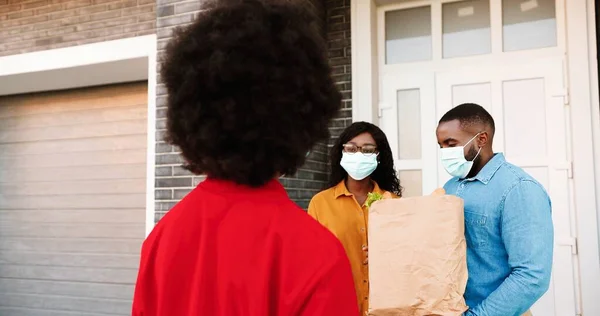 Parte trasera de mensajería femenina afroamericana entrega bolsa con alimentos saludables para parejas en casa, entregando a los hombres al aire libre. Entrega mujer trayendo paquete de verduras al hombre y la mujer. Concepto de Coronavirus —  Fotos de Stock