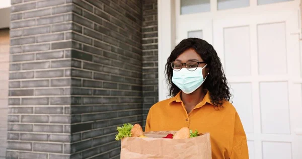 El mensajero femenino afroamericano trasero entrega el bolso con la comida sana al cliente en casa y entrega al aire libre. Entrega mujer trayendo paquete de verduras al cliente. Concepto de Coronavirus. Cuarentena —  Fotos de Stock