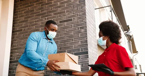 Mensajera afroamericana en máscara entregando paquetes de cartón a un cliente masculino en casa y entregando afuera. Entrega mujer trayendo cajas de pizza al hombre cliente. Concepto de Coronavirus. —  Fotos de Stock