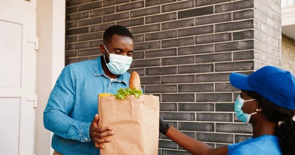 Rear mensajero femenino afroamericano en máscara médica entrega bolsa con comida saludable a la casa del cliente masculino, dando al aire libre y diciendo adiós. Entrega mujer trayendo paquete de verduras al cliente del hombre. —  Fotos de Stock