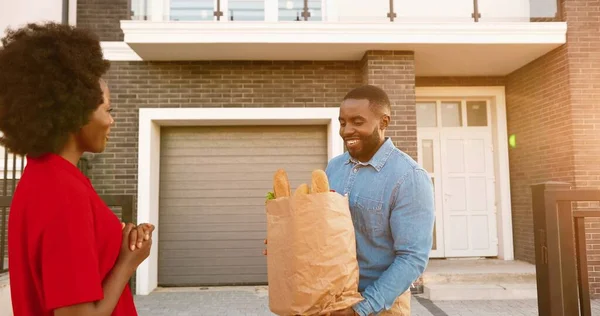 Correio feminino afro-americano entregando saco com comida para cliente masculino em casa, conversando e entregando fora. Mulher de entrega sorrindo e trazendo pacote de produtos para o homem cliente. — Fotografia de Stock