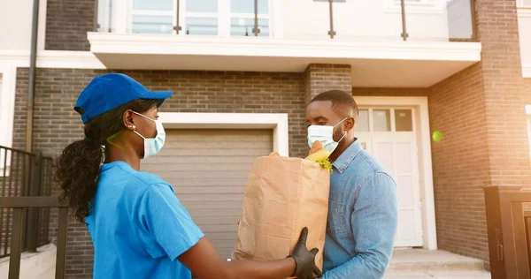 Corriere donna afroamericana in maschera medica che consegna borsa con cibo al cliente maschio a casa e consegna all'esterno. Consegna donna portando pacchetto di prodotti per cliente uomo. Blocco del coronavirus. — Foto Stock