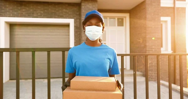 Retrato de la joven mensajera afroamericana en máscara médica, guantes y sombrero de pie en la casa en la calle y la celebración de paquetes de cartón. Trabajadora de partos en uniforme con cajas. Tiempo de cuarentena. —  Fotos de Stock