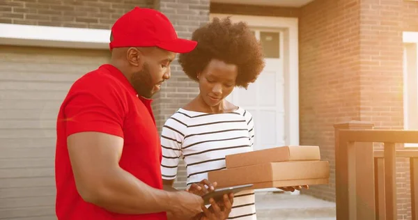 Correio masculino afro-americano em uniforme vermelho e chapéu entregando pacotes de papelão para cliente feminino em casa e entregando ao ar livre. Entrega homem trazendo caixas de pizza para cliente mulher. — Fotografia de Stock