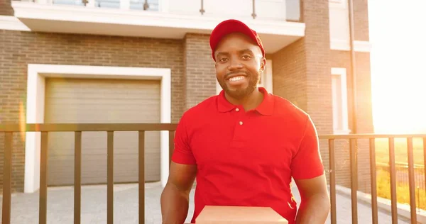 赤い制服を着たアフリカ系アメリカ人の若い男の宅配便の肖像画と帽子が通りに家に立ってカートンの小包を保持します。男性配達労働者は箱と一致している。笑顔で. — ストック写真